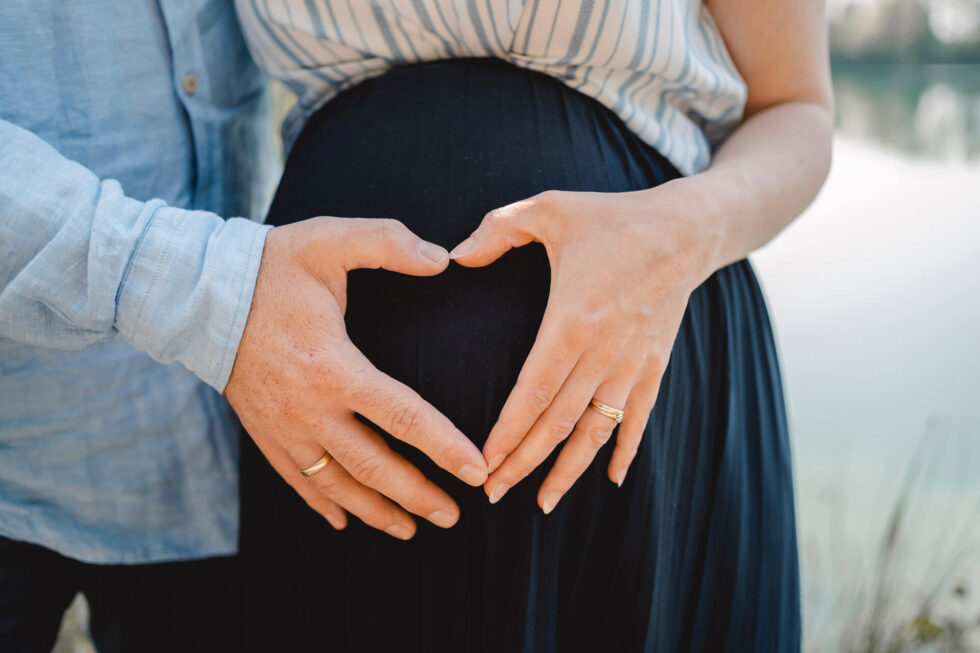 Eine Schwangere und ihr Partner formen mit ihren Händen ein Herz auf dem Babybauch bei einem Babybauch Shooting in München.