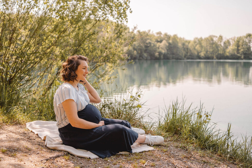 Schwangere Frau sitzt in Gedanken versunken an einem See bei einem Babybauch Shooting in München