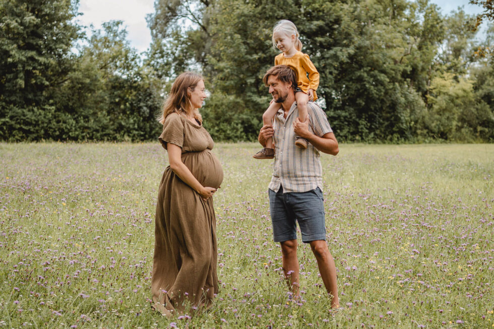Eine Schwangere, die ihren Babybauch hält, und ihrem Partner dabei zusieht, wie er die ältere Tochter in die Luft wirft
