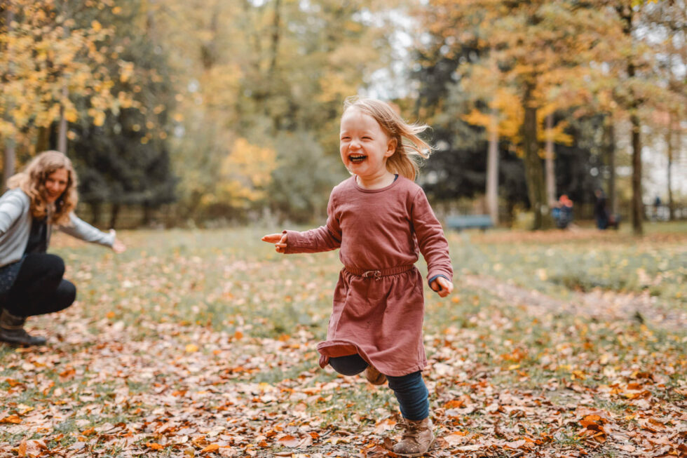 Kleines Mädchen läuft lachend an seiner Mama vorbei, fotografiert bei einem Familien Fotoshooting in München