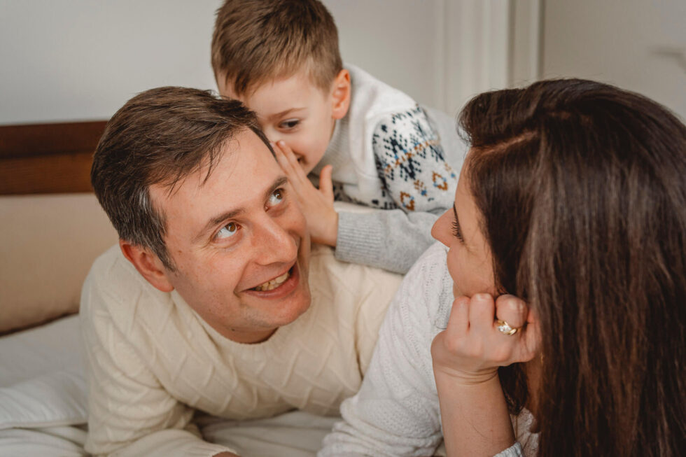 Ein Junge flüstert seinem Vater etwas Lustiges ins Ohr, fotografiert bei einem Familien Fotoshooting in München