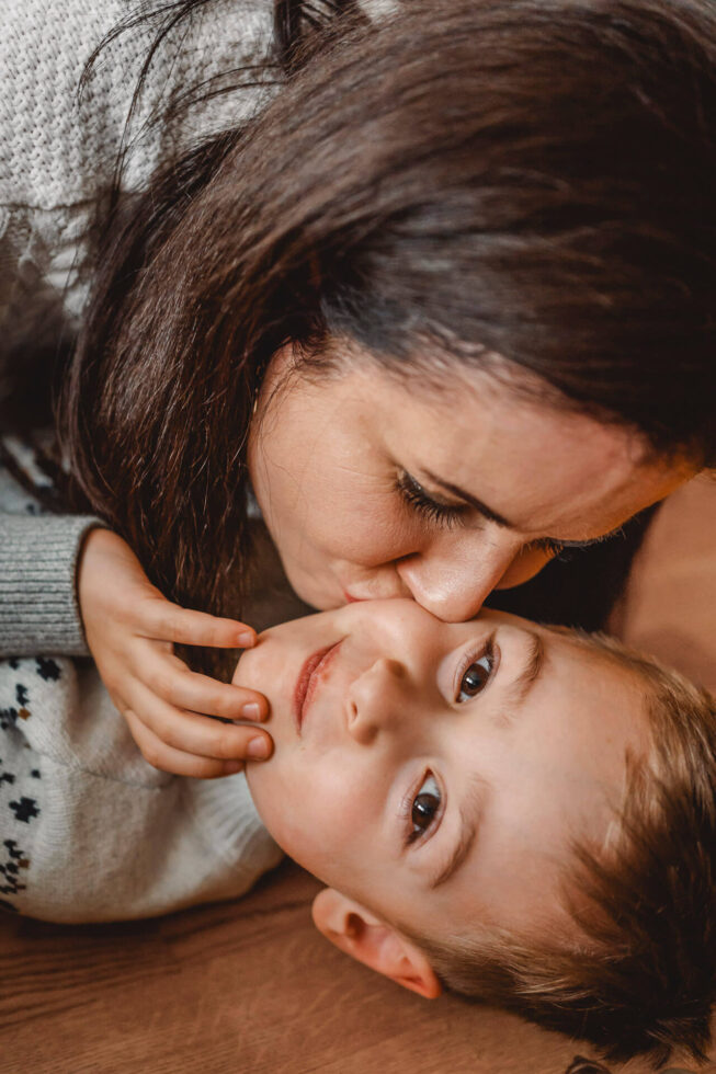 Ein Junge bekommt von seiner Mutter einen dicken Kuss auf die Backe gedrückt, fotografiert bei einem Familienfotoshooting in München
