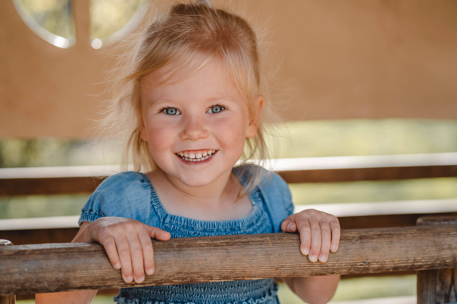 kindergartenfotografie-muenchen-171 Porträt eines Mädchens das sich in einem Spielhaus an einer Querstange festhält und in die Kamera lacht aufgenommen von einem Kindergartenfotograf München