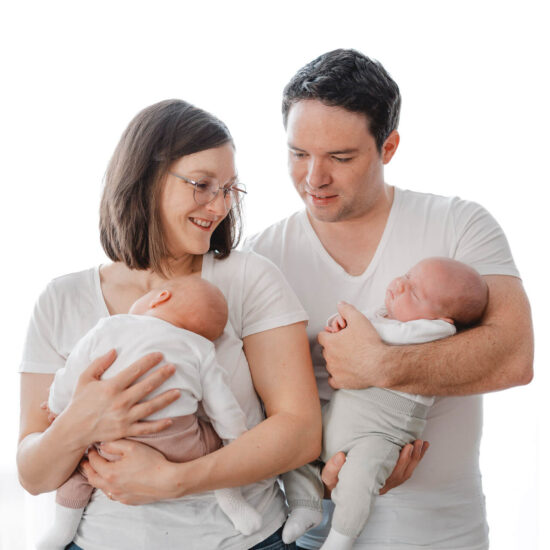 Eine Mutter und ein Vater, die jeweils eins ihrer neugeborenen Zwillinge auf dem Arm halten, fotografiert bei einem Neugeborenen Shooting in München